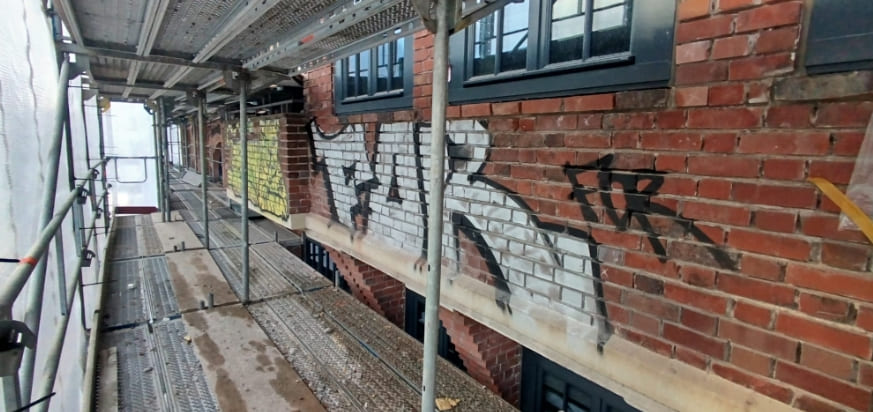 Colorful graffiti tags and pieces on a red brick wall, with metal scaffolding indicating access for maintenance or removal.
